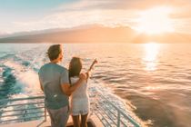 A couple stands on a boat deck at sunset, looking out toward a distant coastline as the vessel leaves a wake.