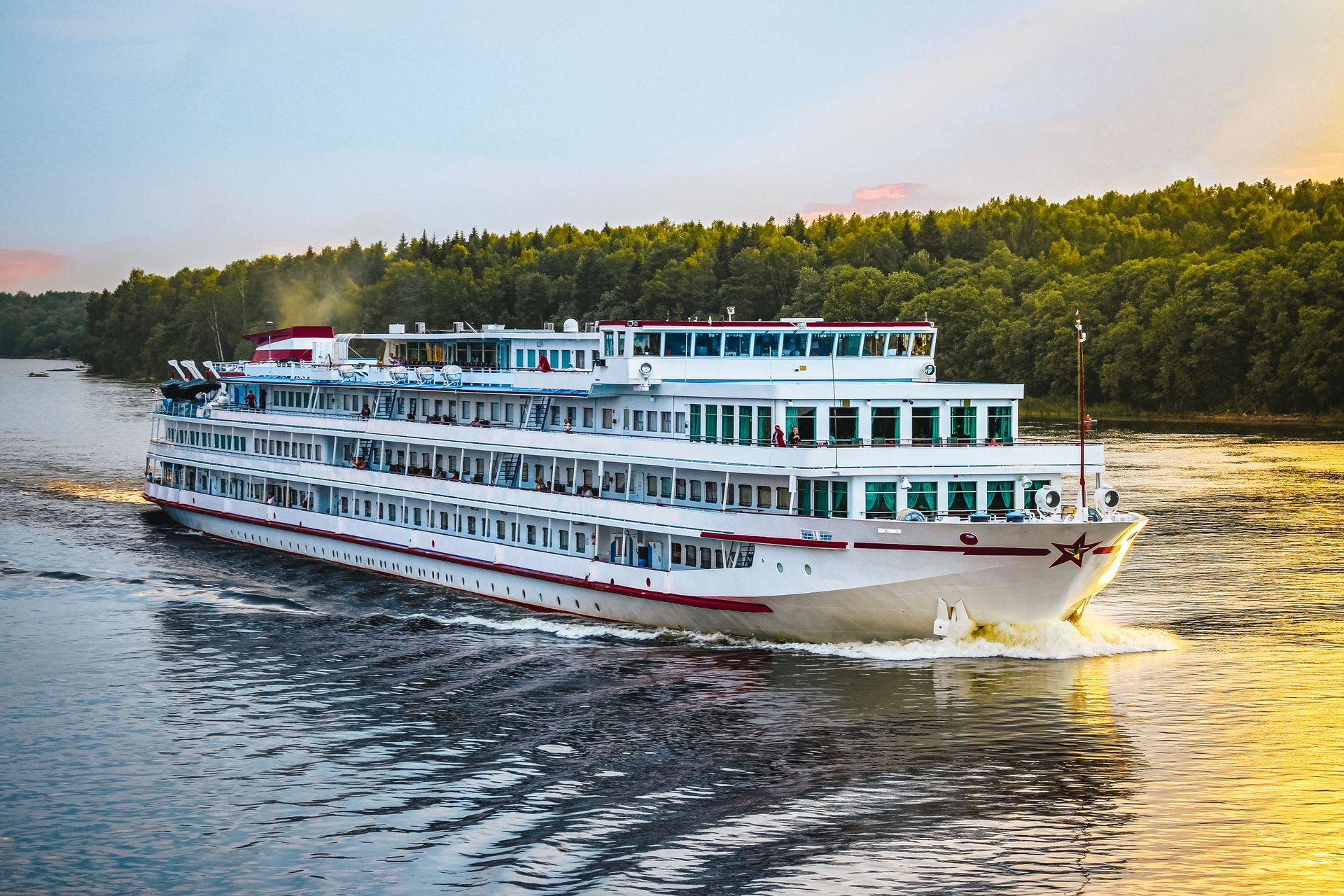 A white river cruise ship travels along a calm waterway lined with green trees under a warm, golden sunset sky. A white river cruise ship travels along a calm waterway lined with green trees under a warm, golden sunset sky.