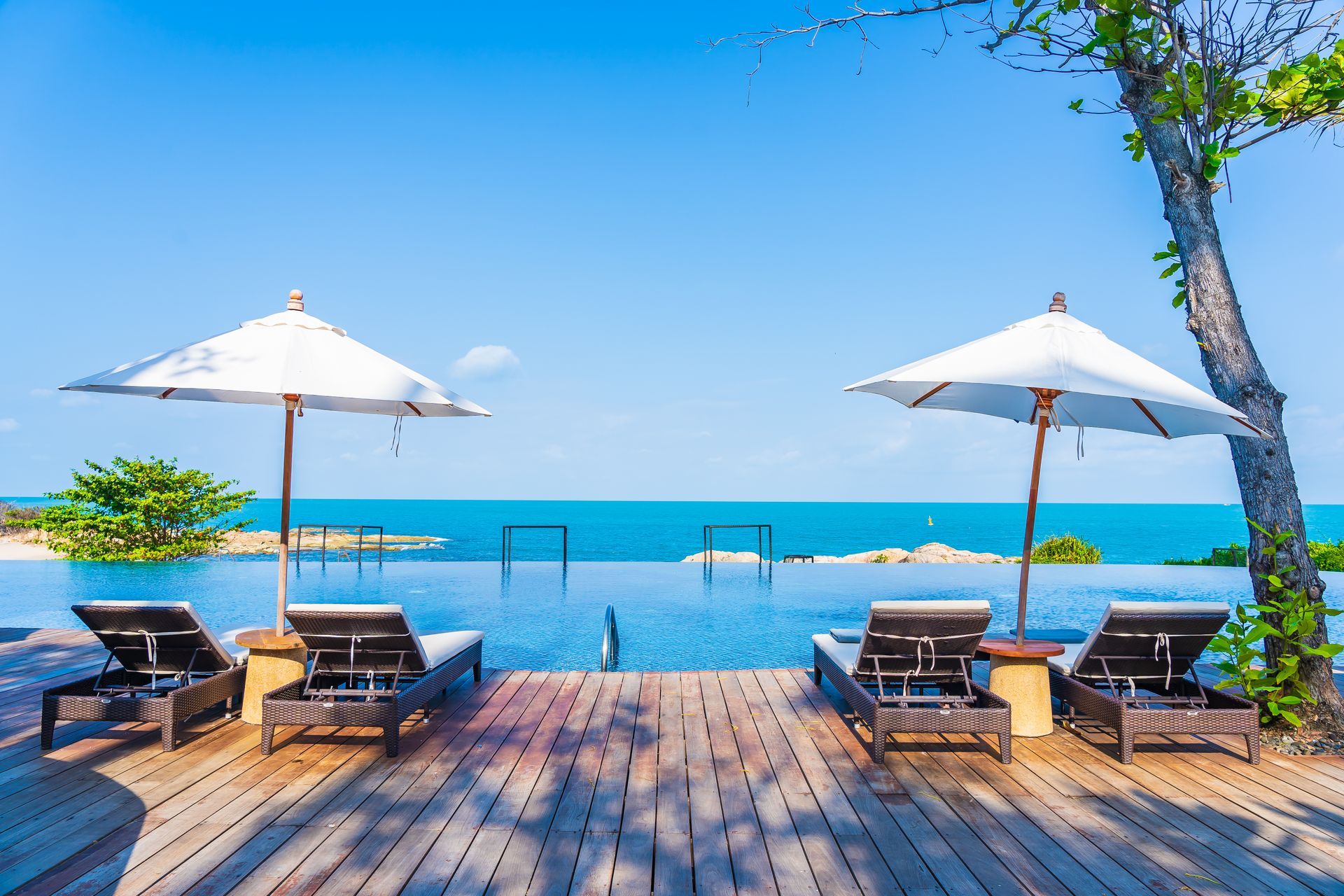 Four sun loungers with two umbrellas on a wooden deck overlooking a blue infinity pool and the sea under a clear sky.