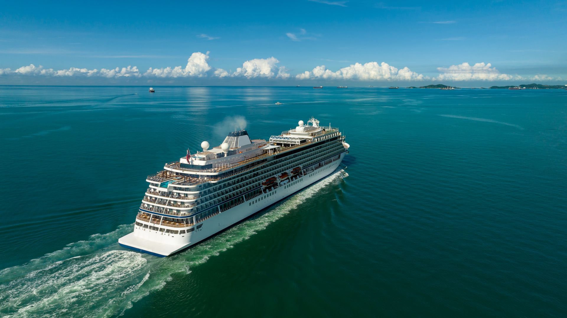 A large white cruise ship sails across a vibrant blue ocean under a bright, sunny sky with scattered clouds.