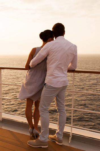 A couple stands at the railing of a cruise ship, watching the sunset over the ocean.