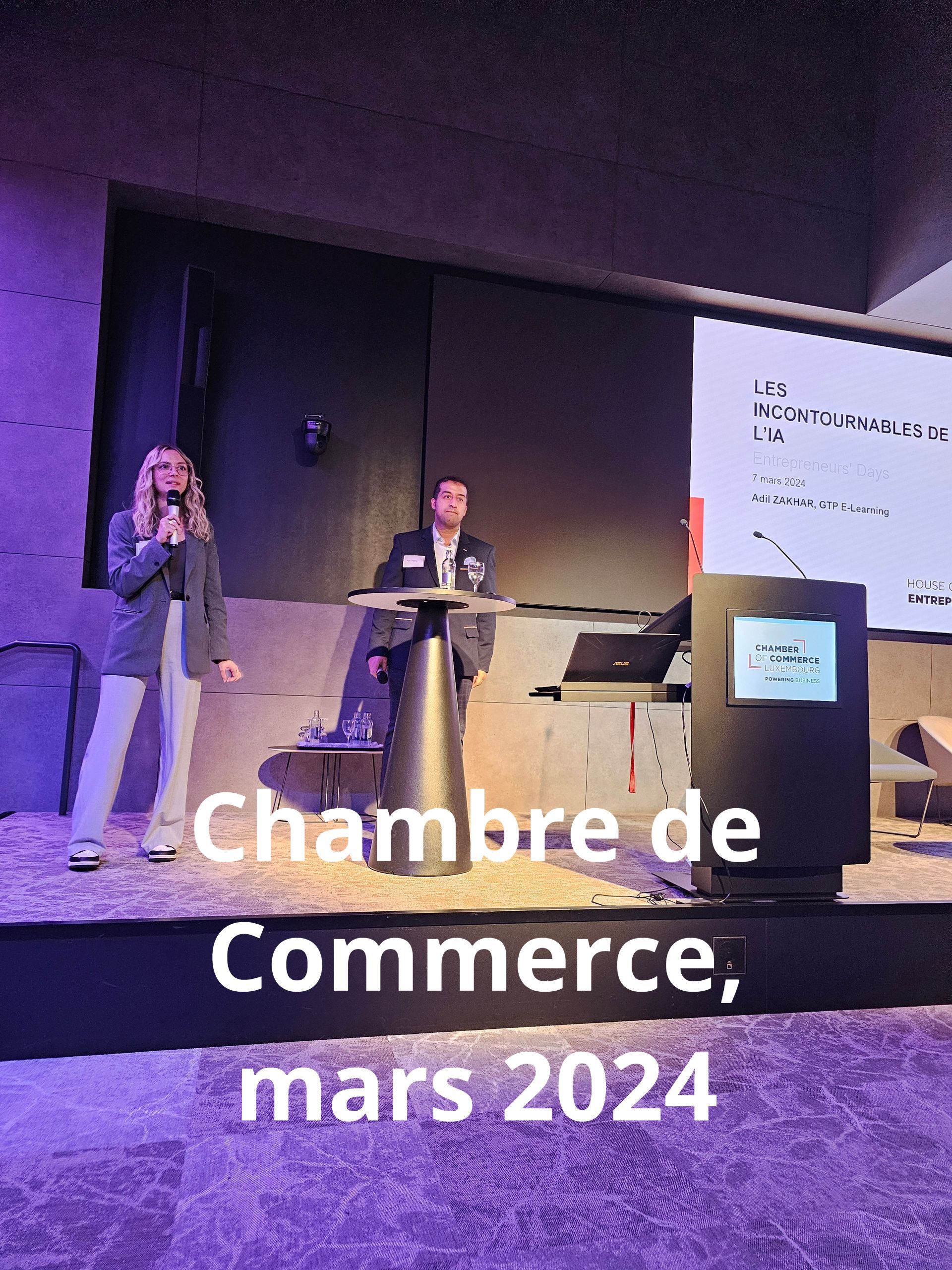 Deux personnes lors d'un événement de la Chambre de commerce, mars 2024. Une femme parle à un podium, un homme est assis à une table avec un ordinateur portable.