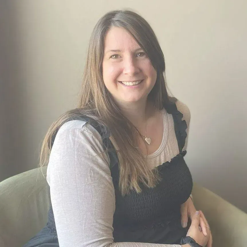 A smiling person with long brown hair, wearing a long-sleeved cream shirt under a black dress, sitting in a chair.