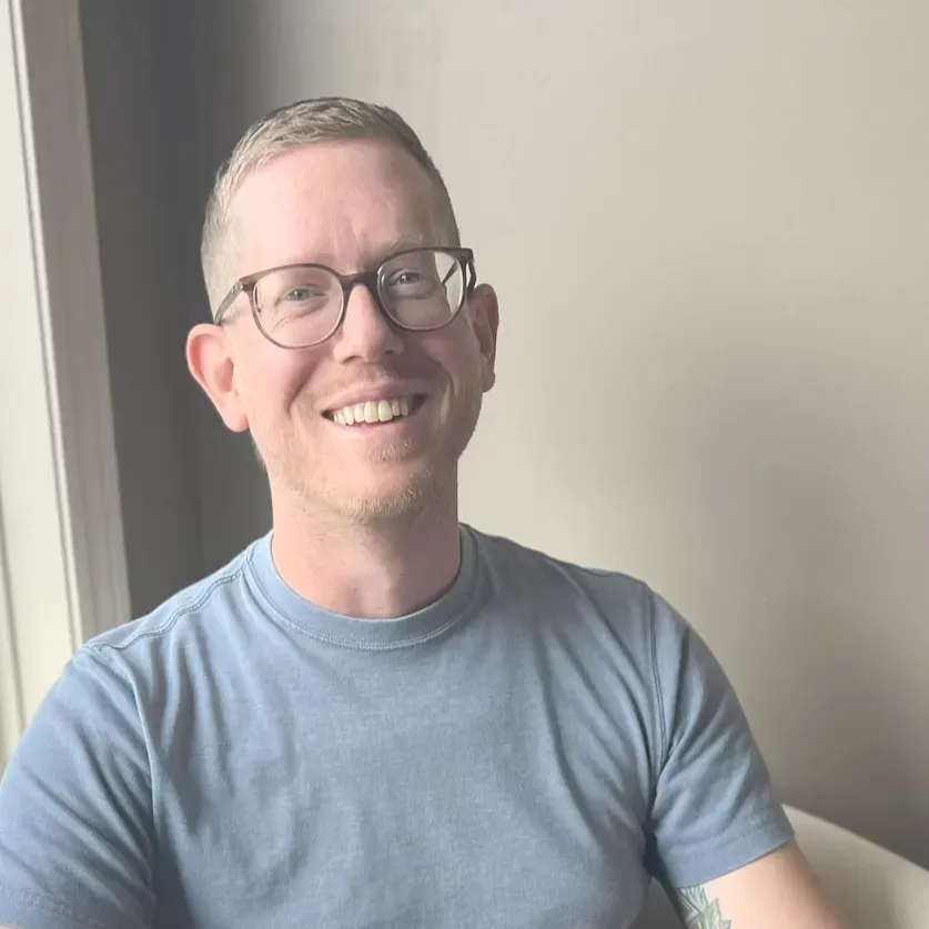A smiling person wearing glasses and a blue t-shirt, posed indoors against a neutral, plain wall.