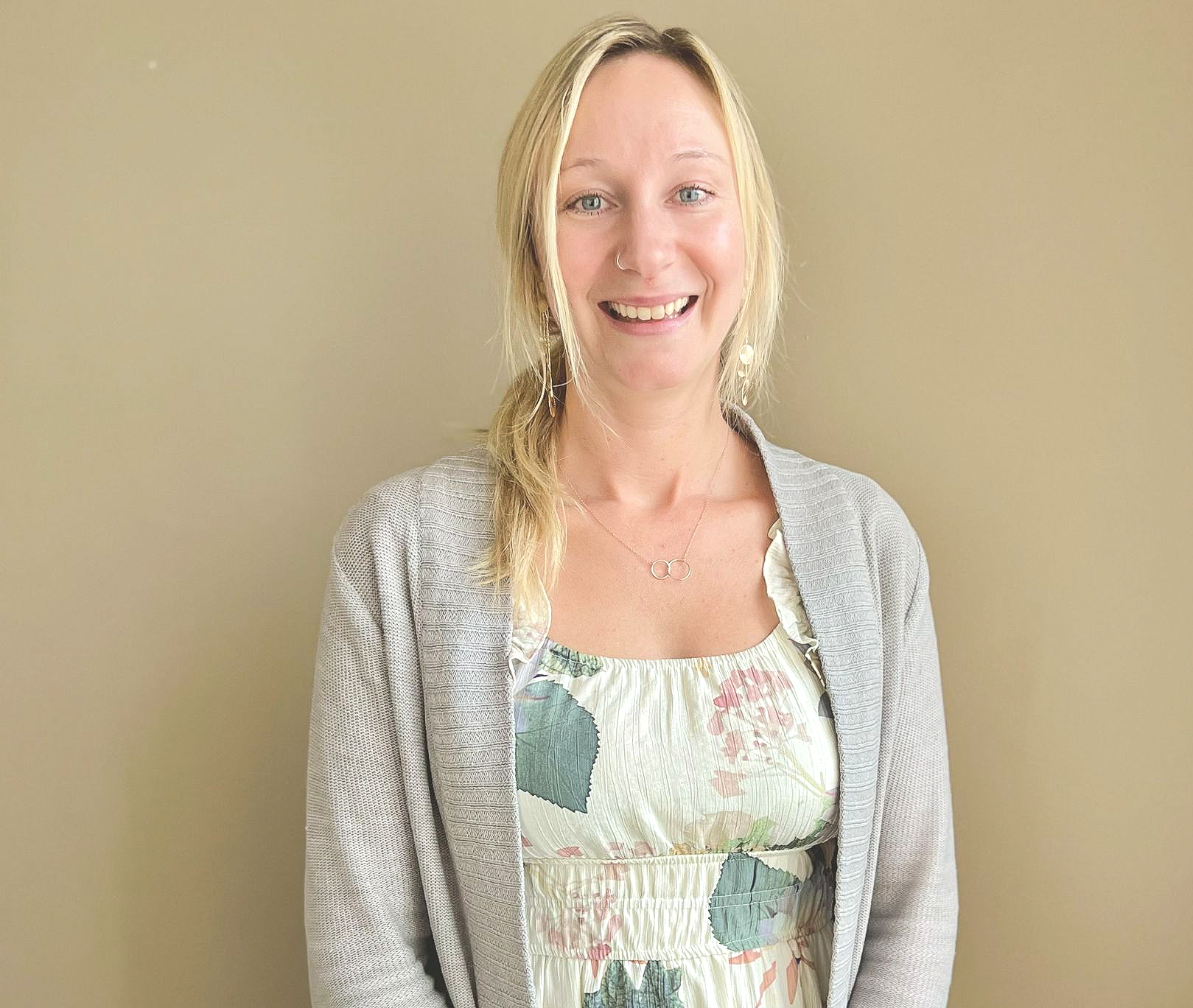 Woman with blonde hair smiles, wearing a floral dress and gray cardigan, standing against a beige wall.