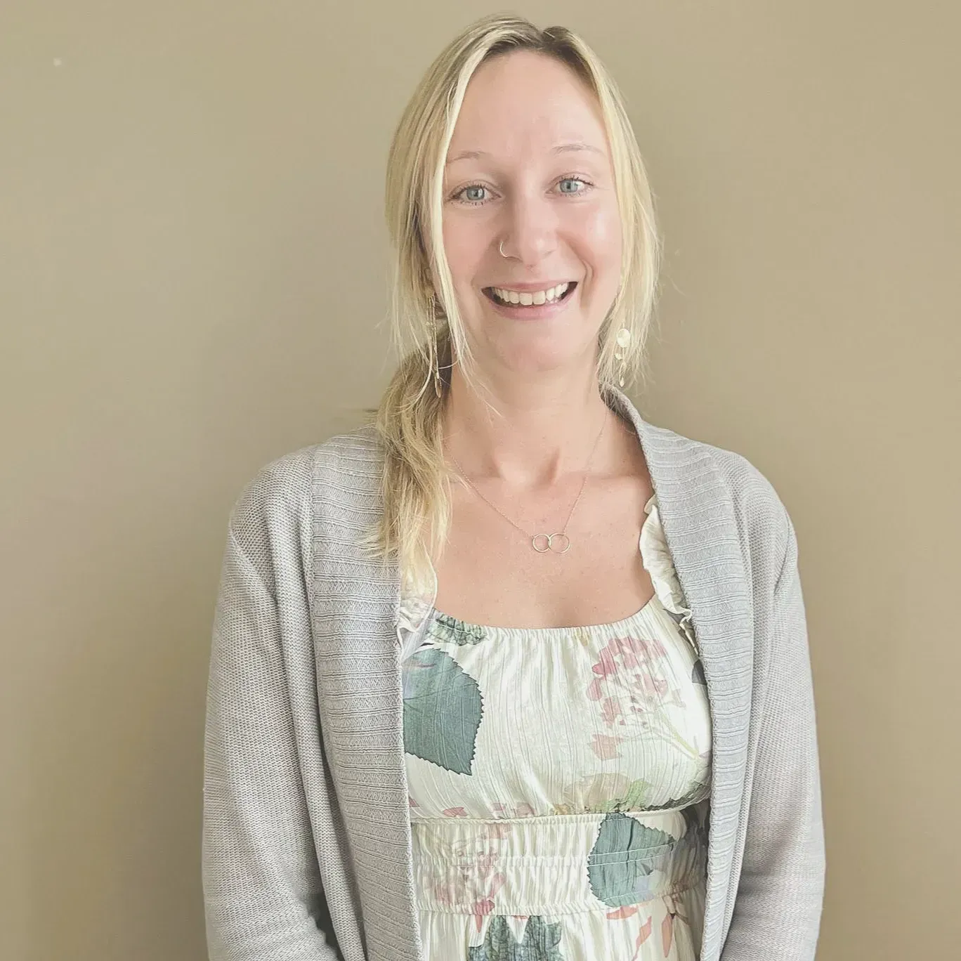 A smiling person with blonde hair in a low ponytail, wearing a grey cardigan over a floral print dress.