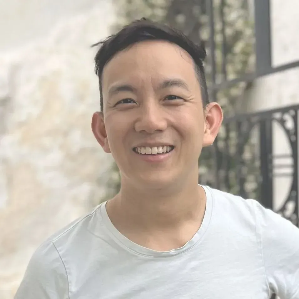 A smiling person with dark hair wears a white t-shirt, posed outdoors against a blurred stone wall and metal gate.