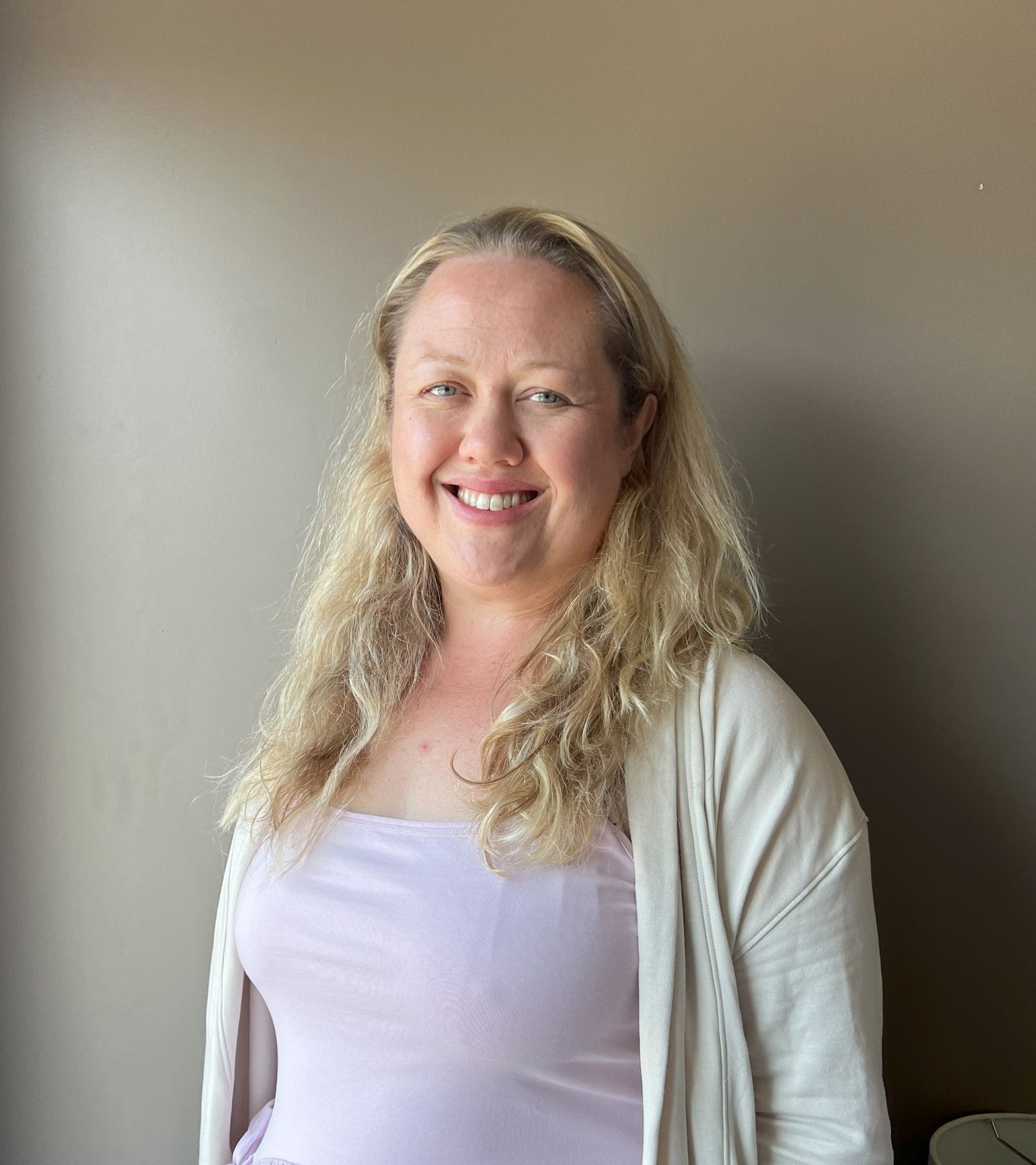 Woman with blonde wavy hair smiles, wearing a pink top and cream cardigan, standing near a beige wall.