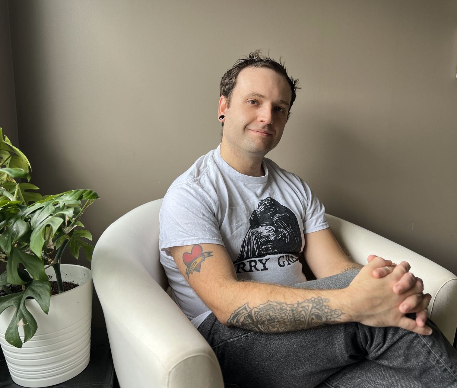 Man sitting in a white chair near a potted plant. He has a tattoo and is wearing a gray shirt, smiling.