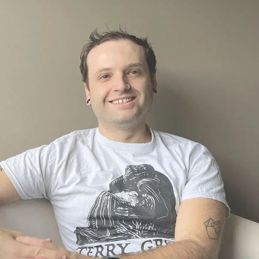 A smiling person with a nose ring and gauges, wearing a white t-shirt with a graphic print, sitting against a beige wall.