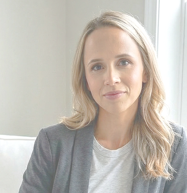 Woman with blonde hair smiling, wearing a pink top and cream cardigan, standing against a beige wall.