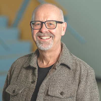 A smiling person with a goatee and glasses wearing a textured gray button-up shirt over a black shirt.