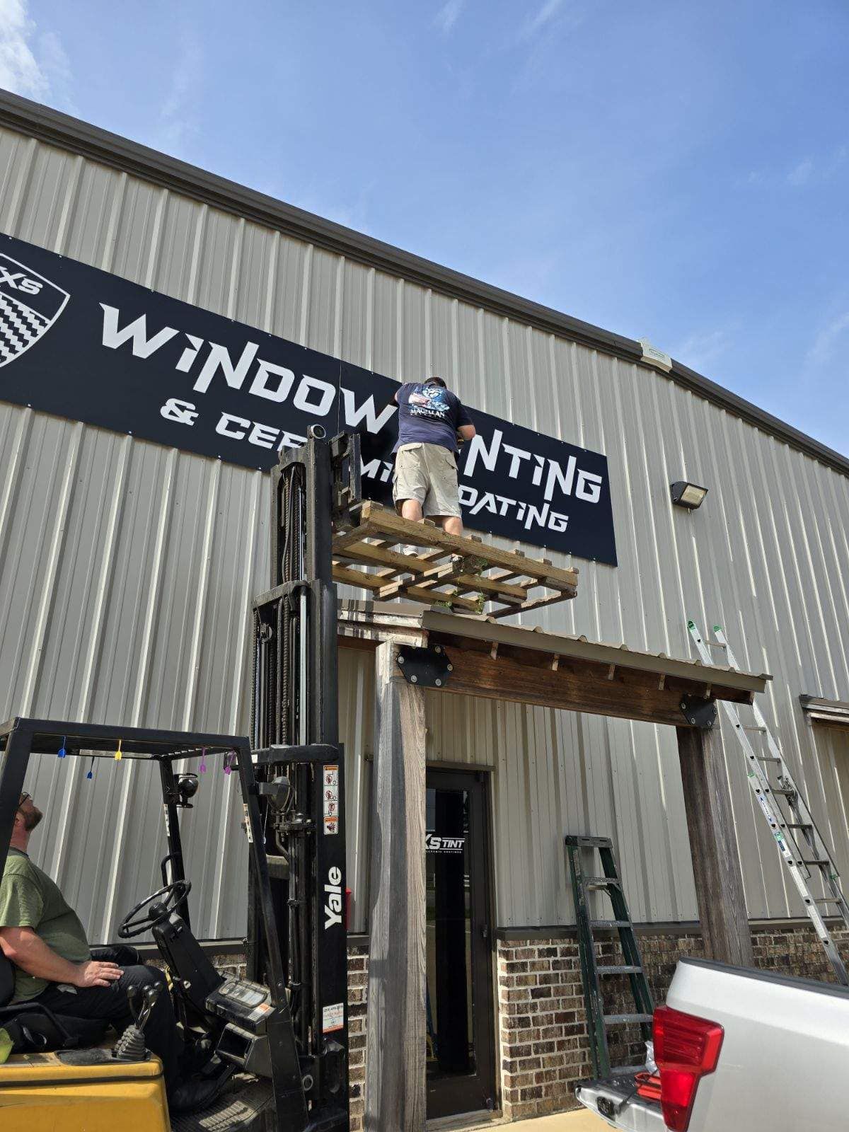 A man is standing on top of a forklift hanging a sign on the side of a building.