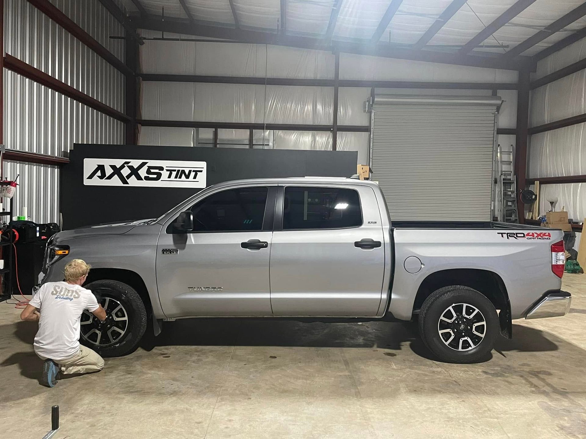 A man is kneeling next to a silver truck in a garage.
