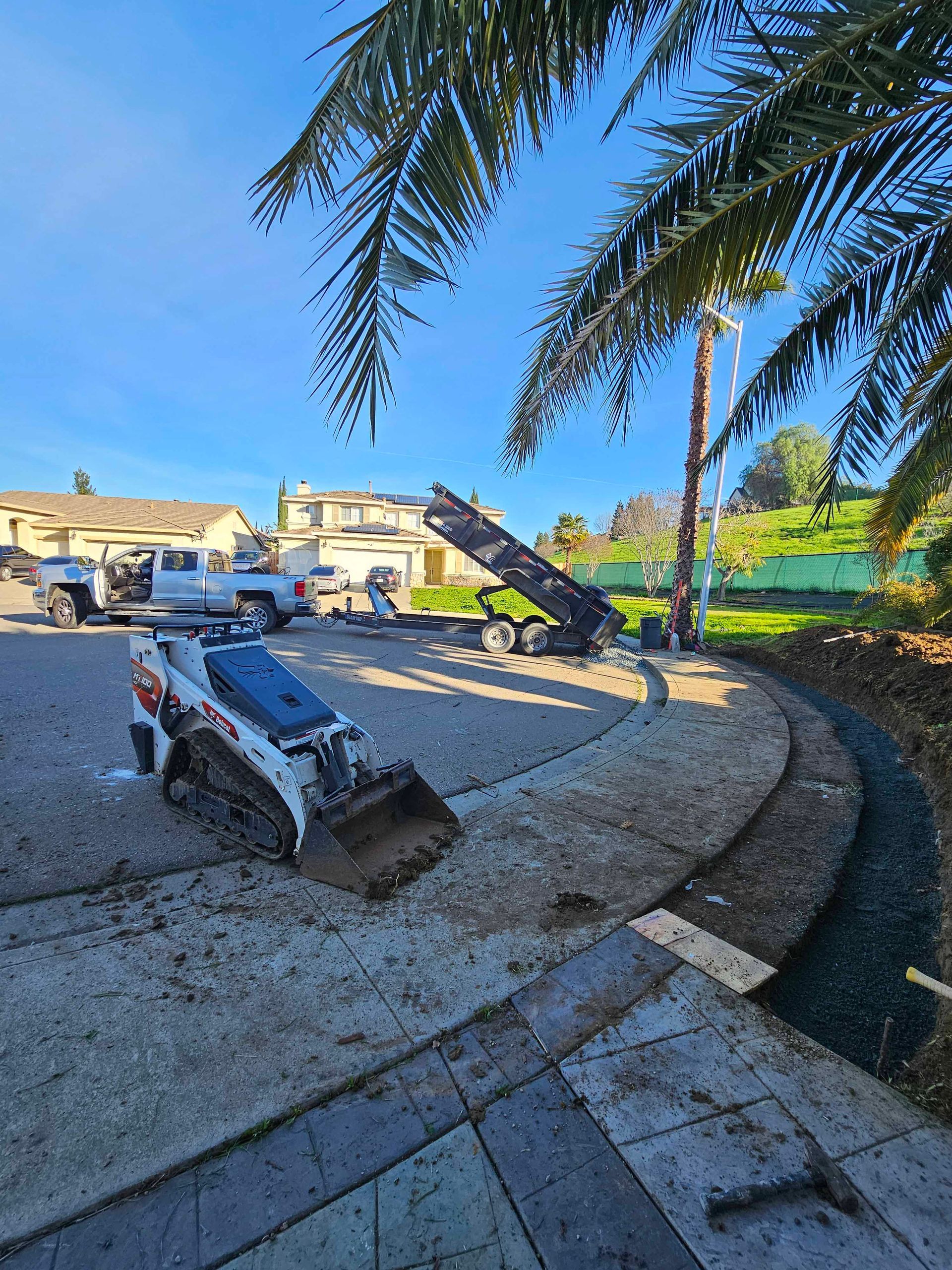 A Bobcat skid steer and truck unloading gravel at a construction site with a palm tree.