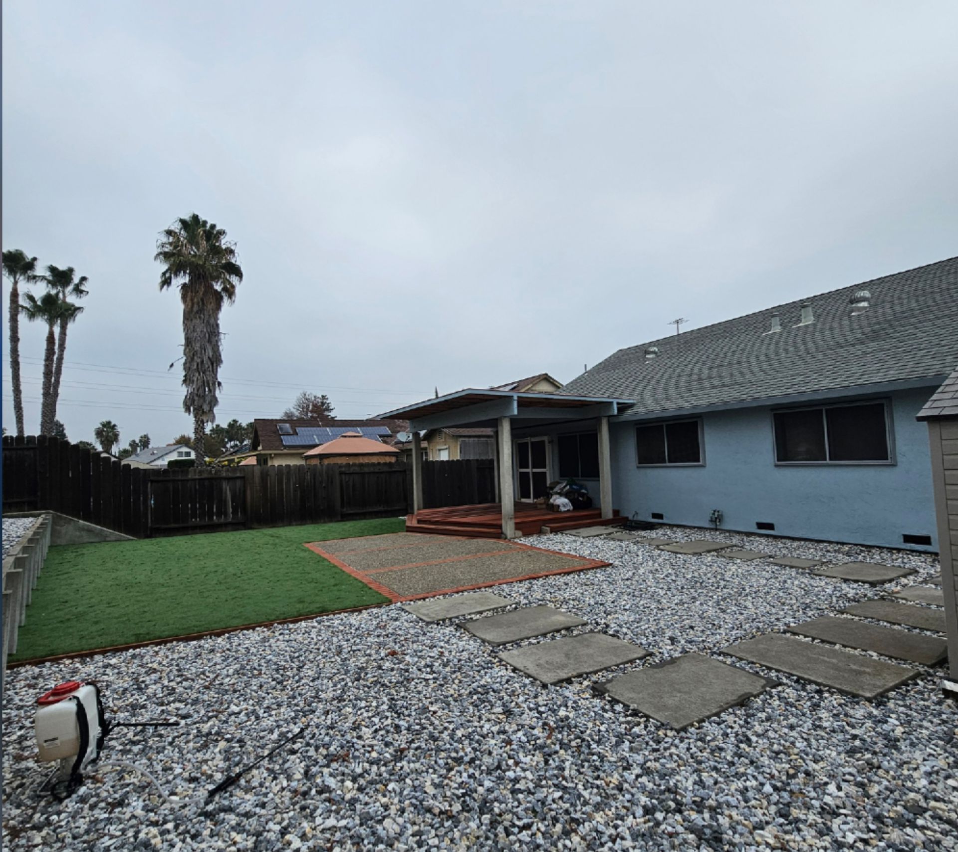 Backyard with gray gravel, concrete stepping stones, artificial turf, a palm tree, and a light blue house.