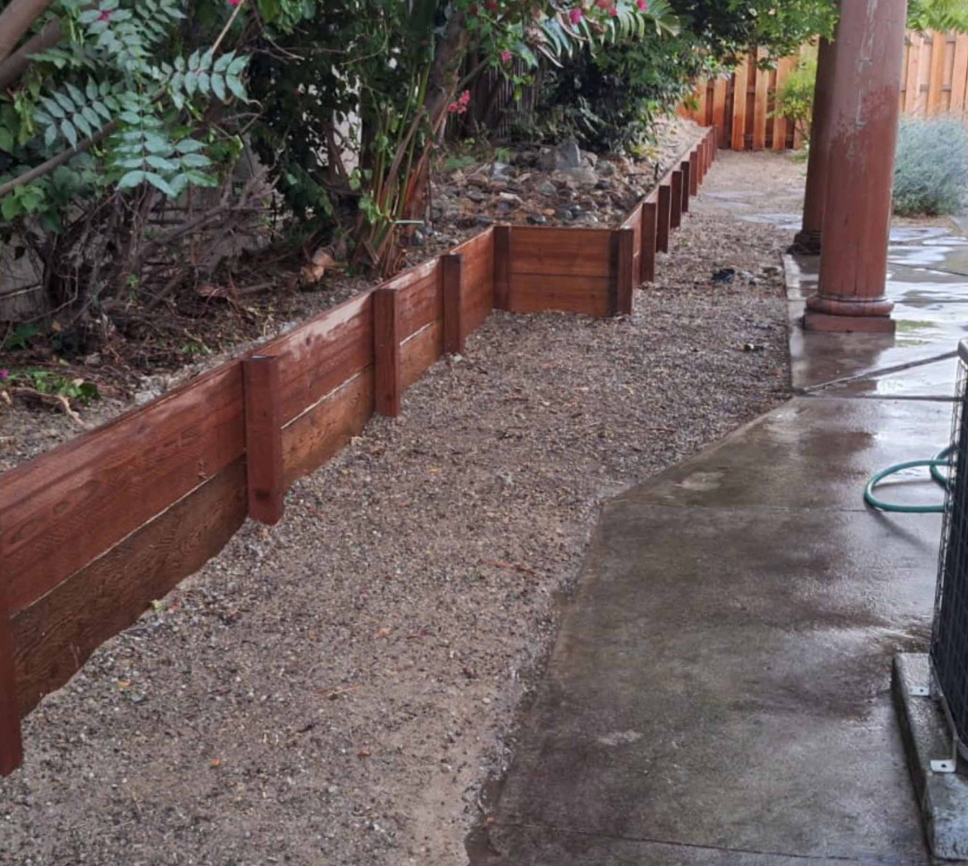 Wooden retaining wall filled with gravel next to a concrete path and garden.