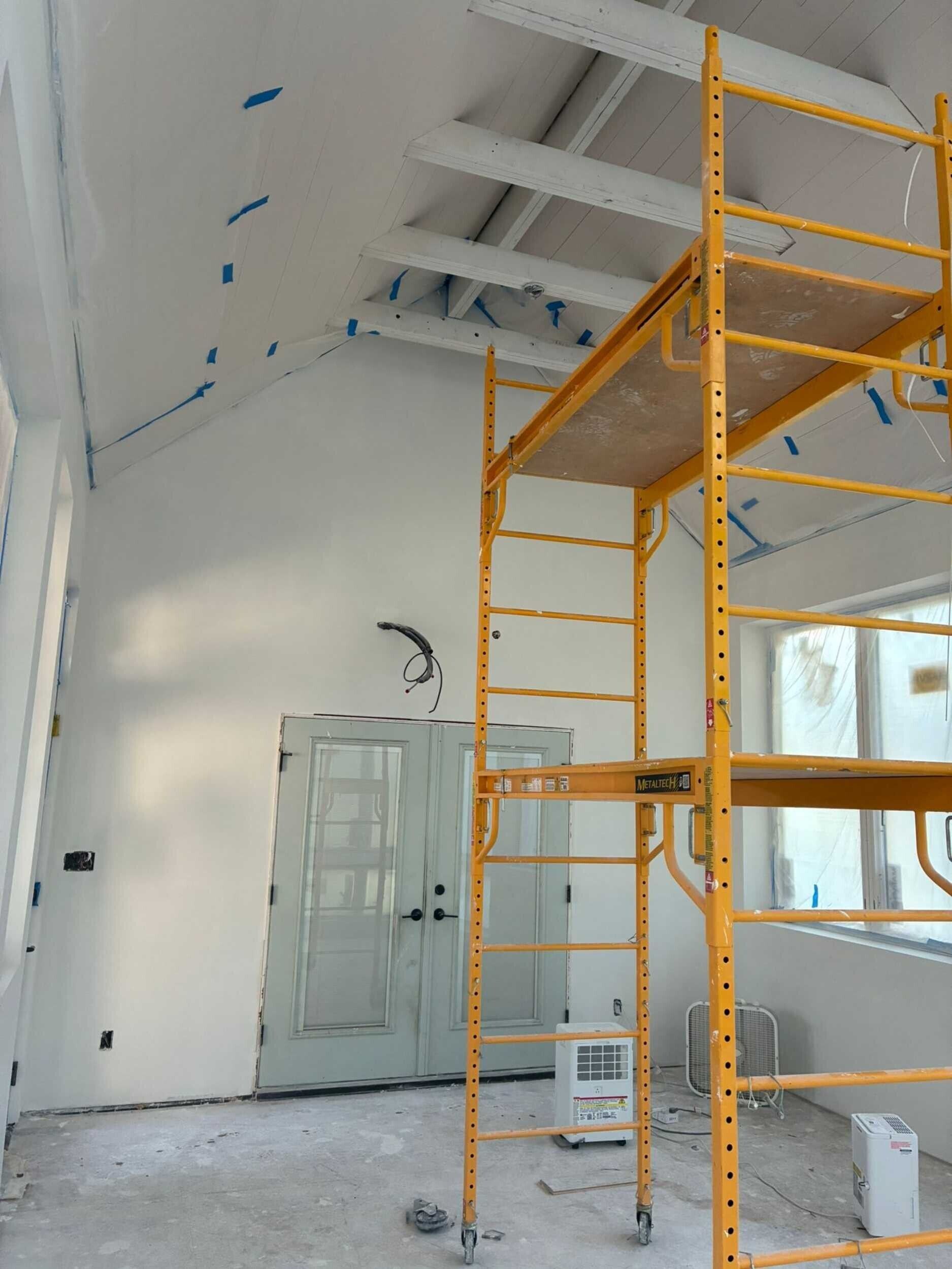 Interior construction: vaulted ceiling with exposed beams, scaffolding, door, and white walls.