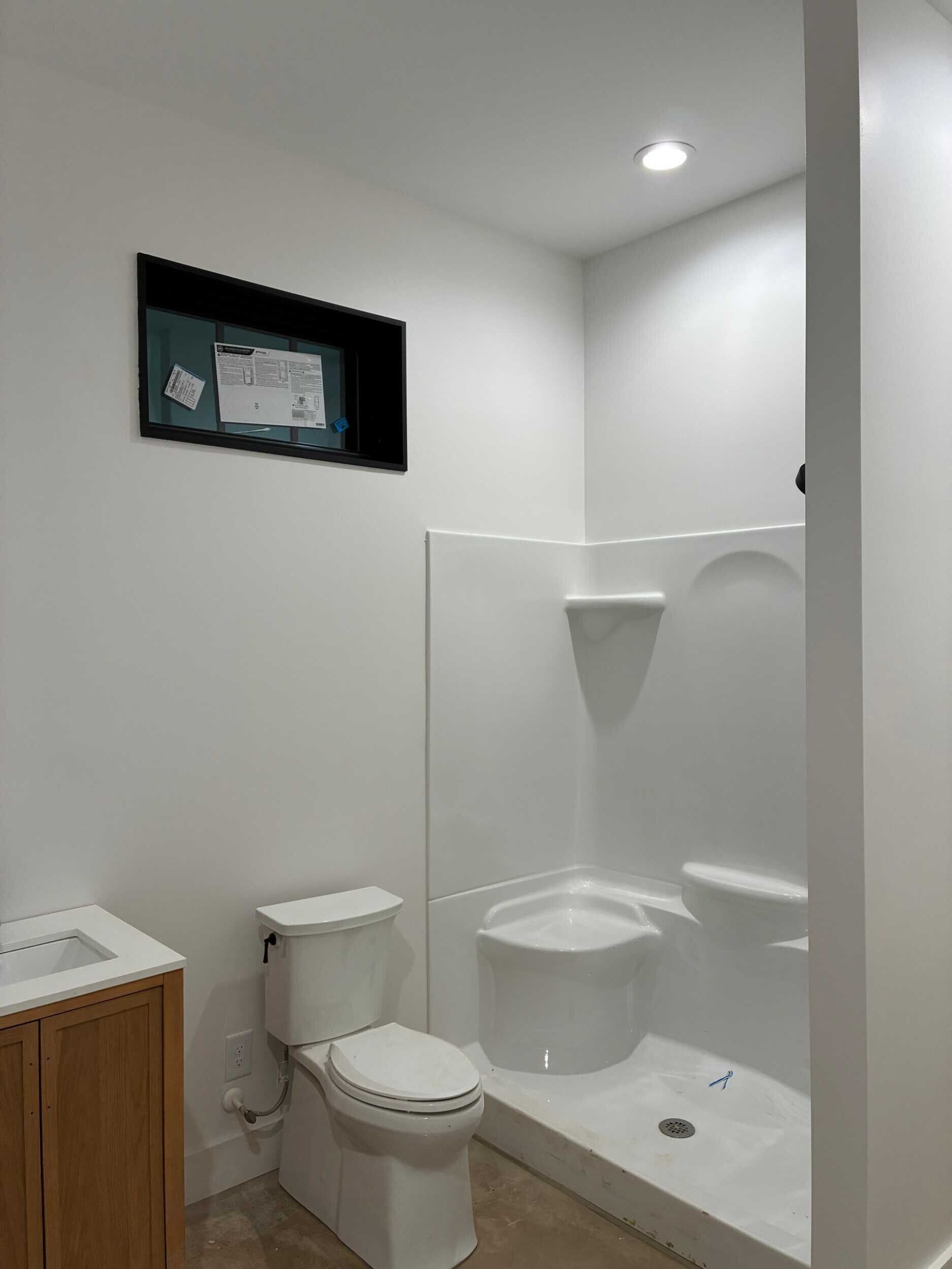 Small, white bathroom with a corner shower, toilet, and vanity. A small window is above the toilet.