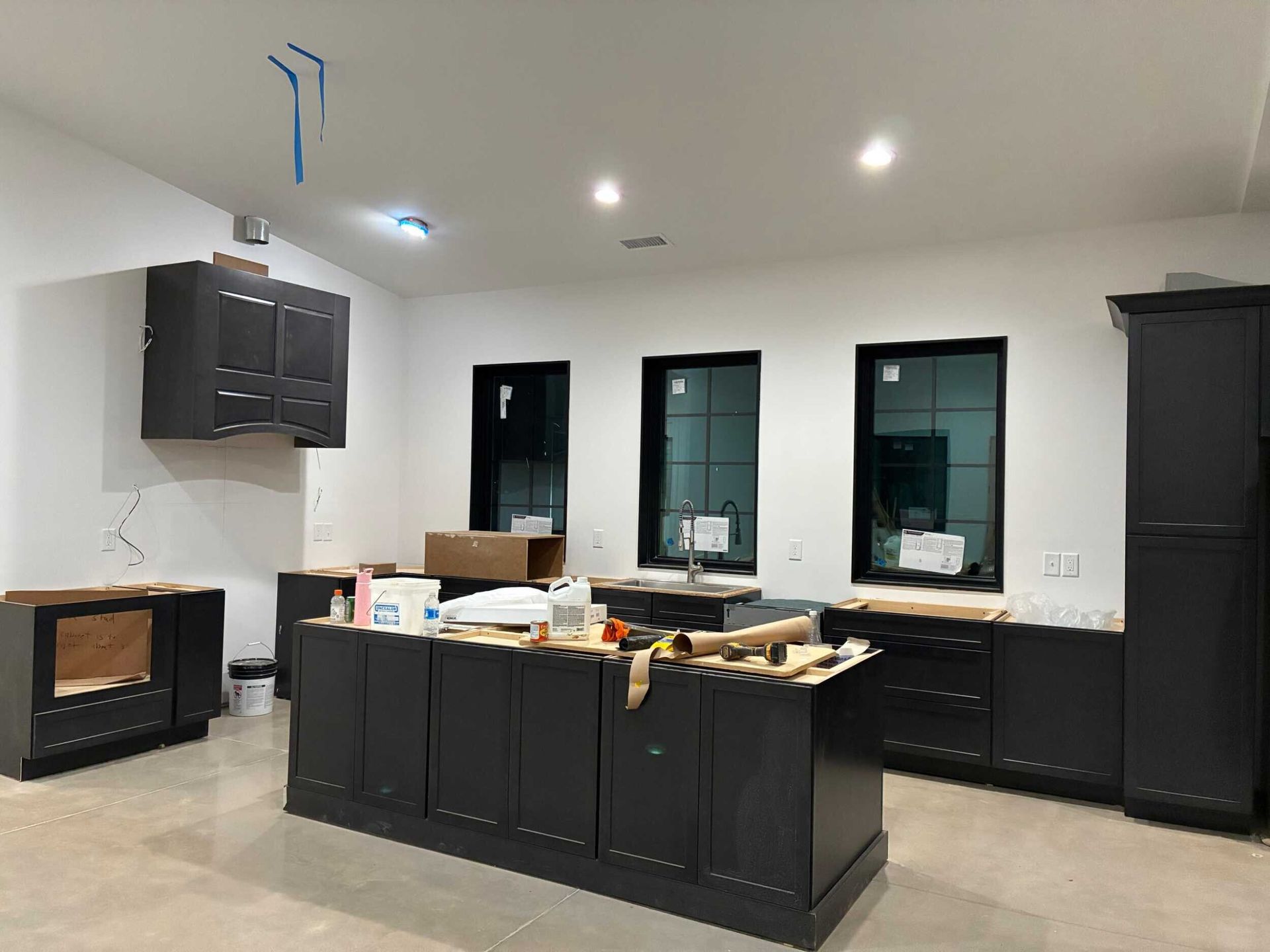 Kitchen under construction with dark cabinets, white walls, and black-framed windows.