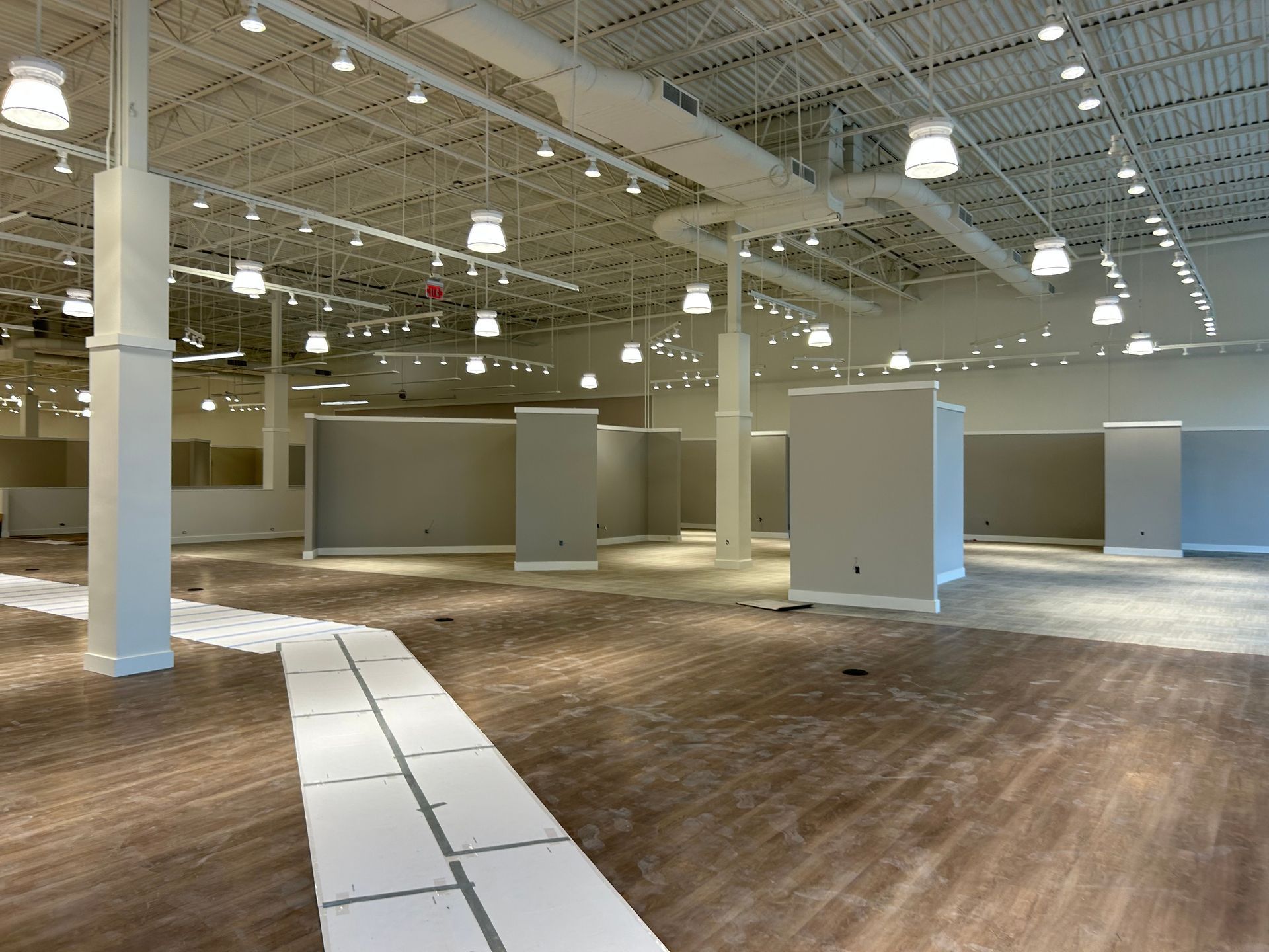 Empty retail space with white walls, ceiling, and columns. Gray partitions on wood floor. Overhead lighting.