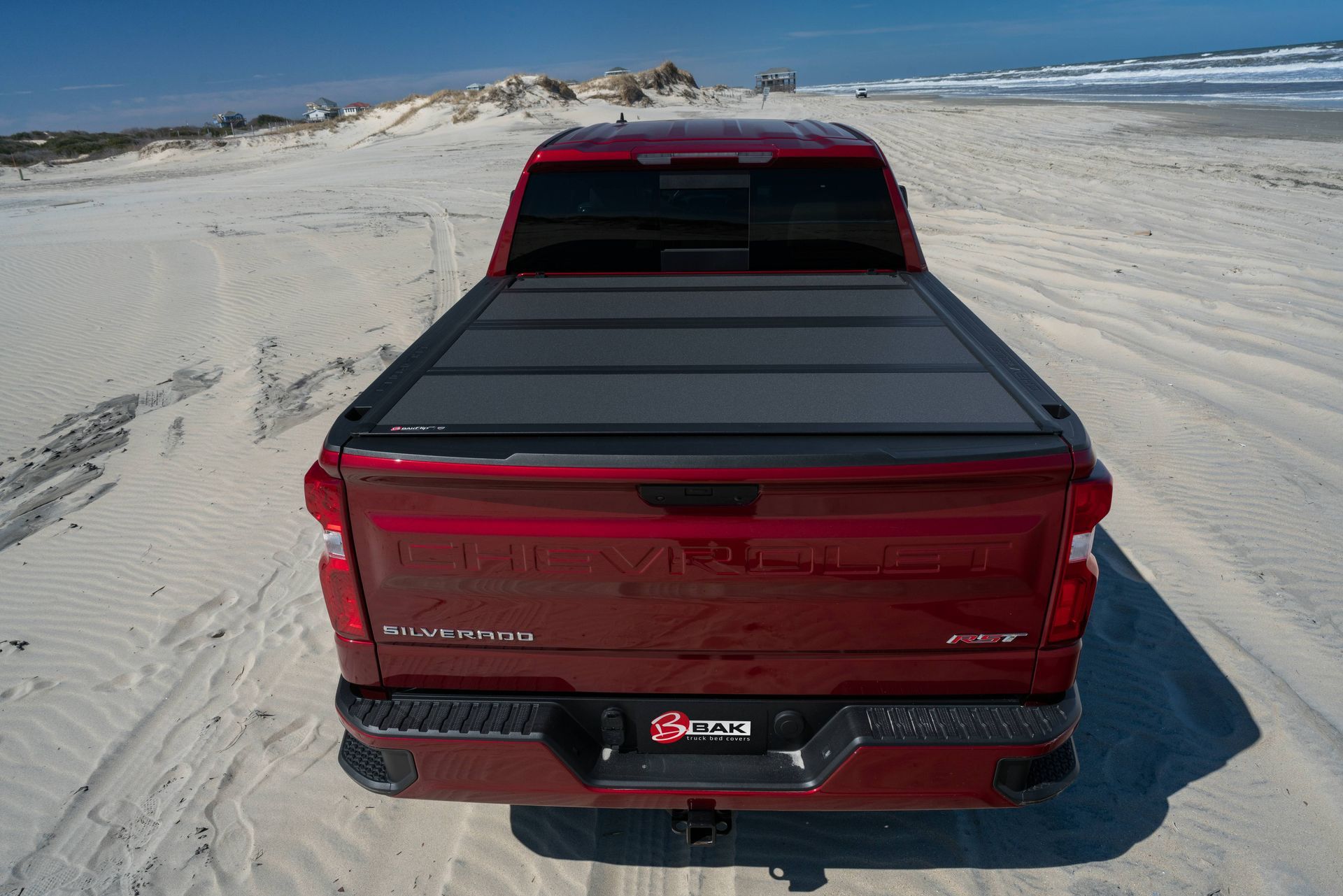 A red truck with a black bed cover is parked on a sandy beach.