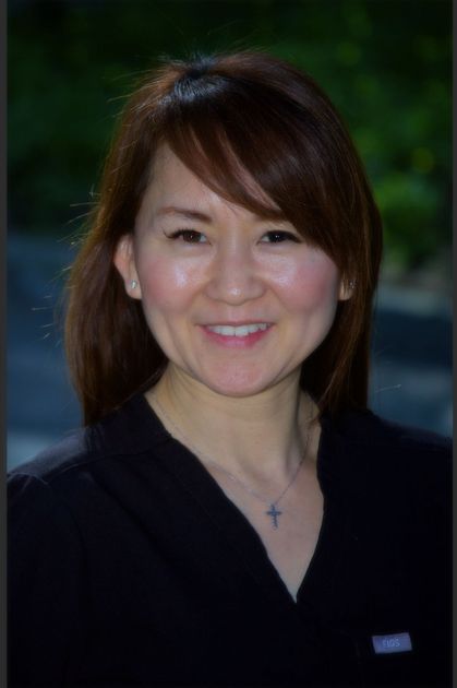 A smiling person with shoulder-length brown hair, wearing a black top with a small cross necklace against a dark background.