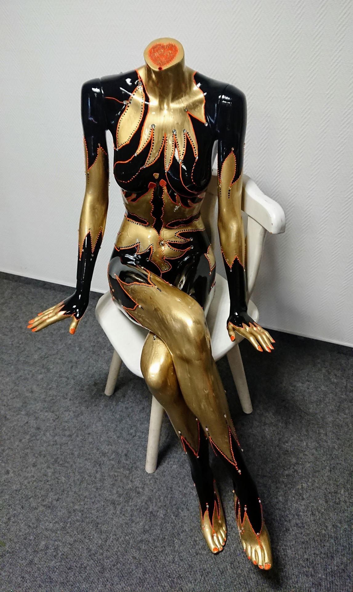 Eine schwarz-goldene Schaufensterpuppe sitzt auf einem Stuhl.