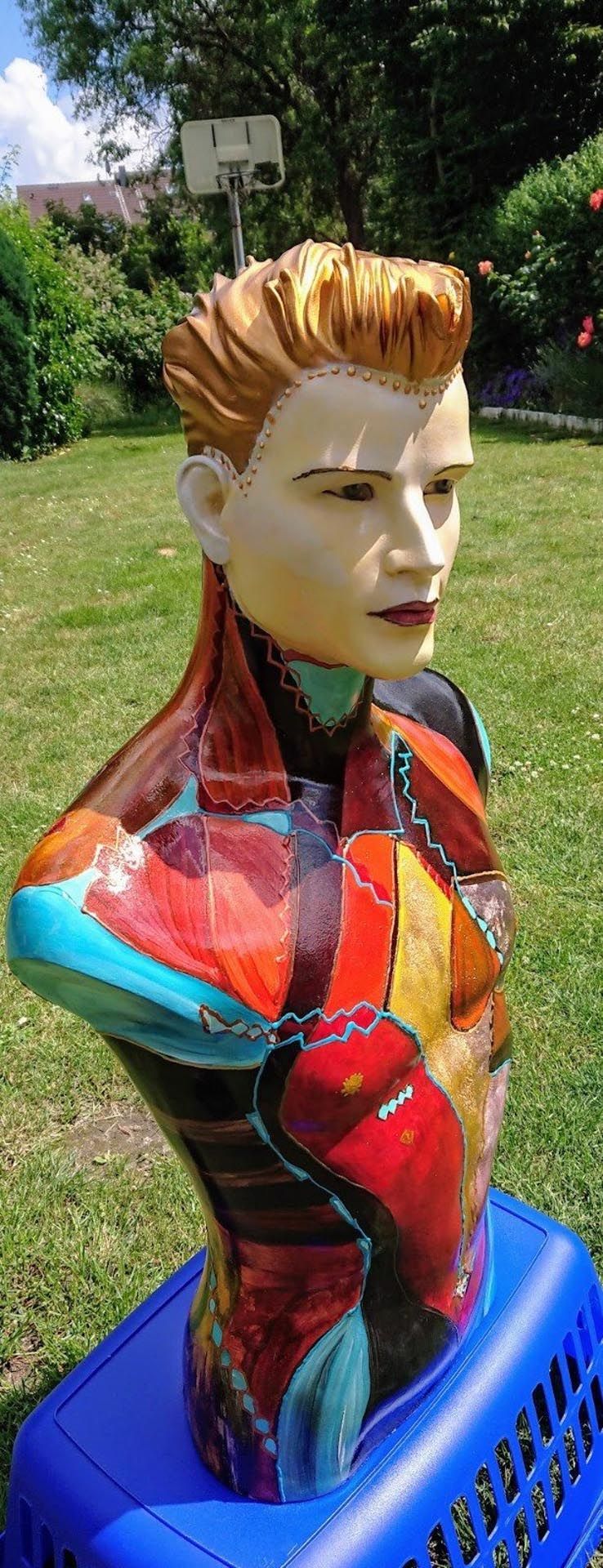 Eine farbenfrohe Statue eines Männertorsos sitzt auf einem blauen Wäschekorb.