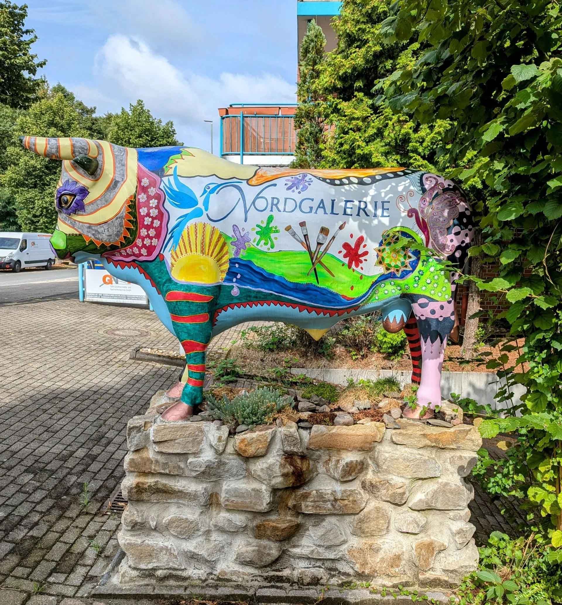 Eine farbenfrohe Kuhstatue sitzt auf einer Steinmauer.