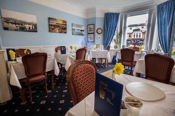 Interior of a restaurant with tables set for dining. Blue walls, patterned carpet, and framed art decorate the space; sunlight streams in the window.
