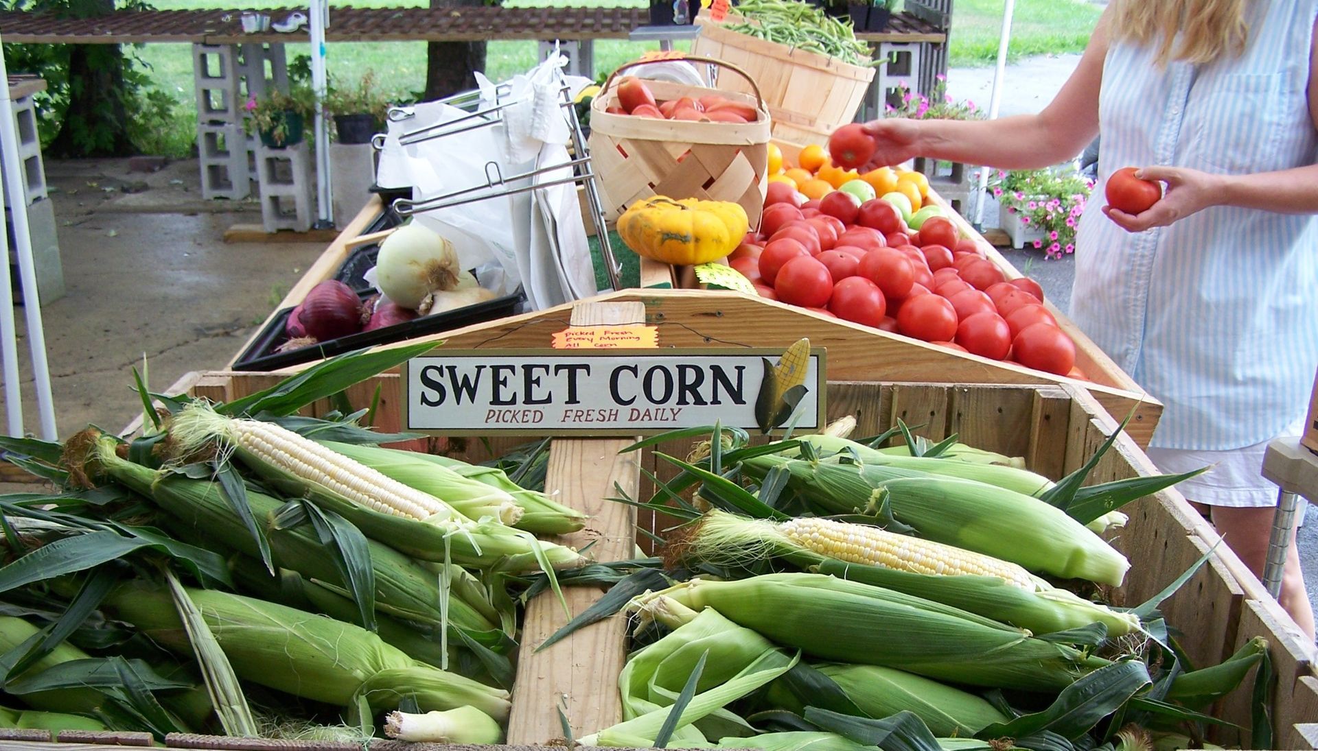 A woman is holding a tomato in front of a sign that says sweet corn