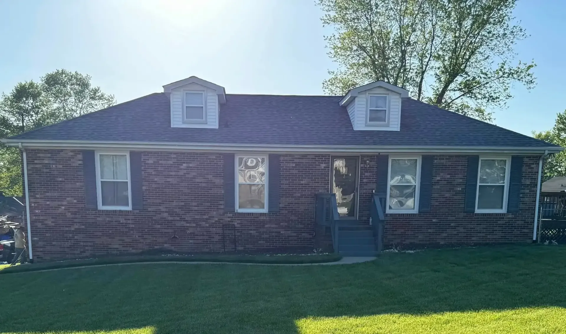 Brick ranch-style house with green shutters, two dormers, and a green lawn on a sunny day.