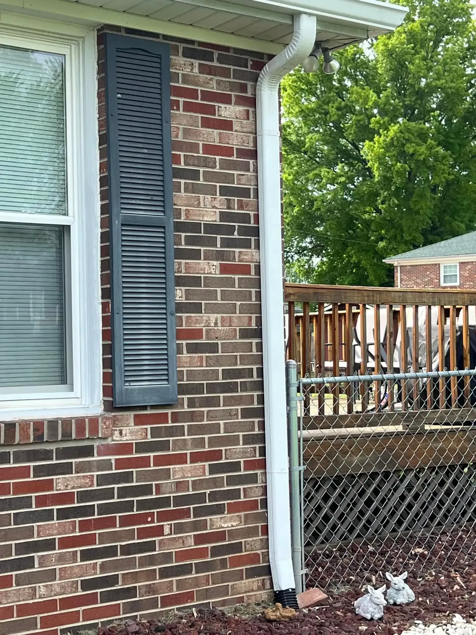 Brick building with white gutter and gray shutter next to a window, with a deck and trees in background.