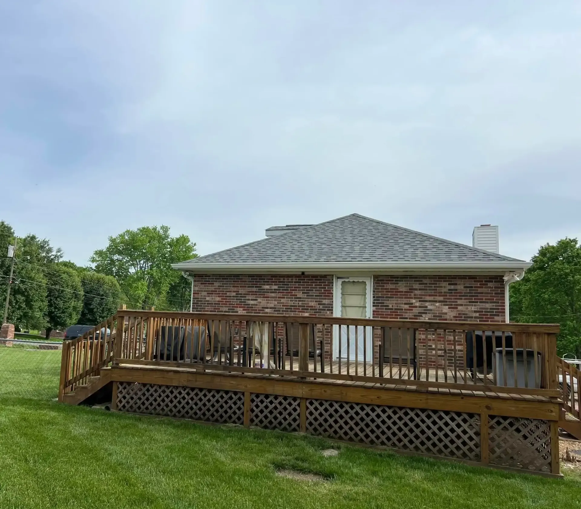 A wooden deck in front of a brick house, with a green yard and trees under a cloudy sky.