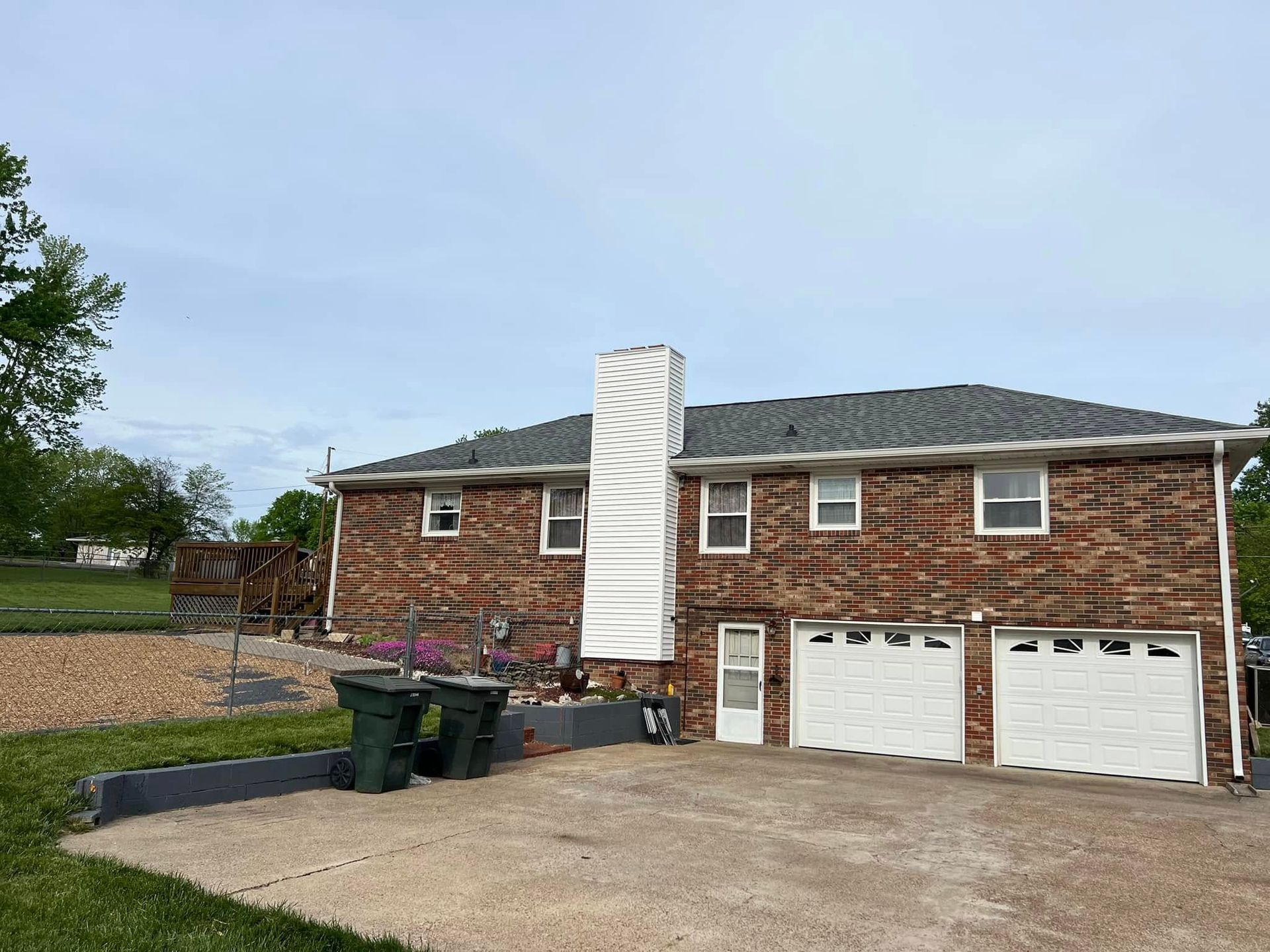 Red brick house with two-car garage, a tall white chimney, and two green trash cans.