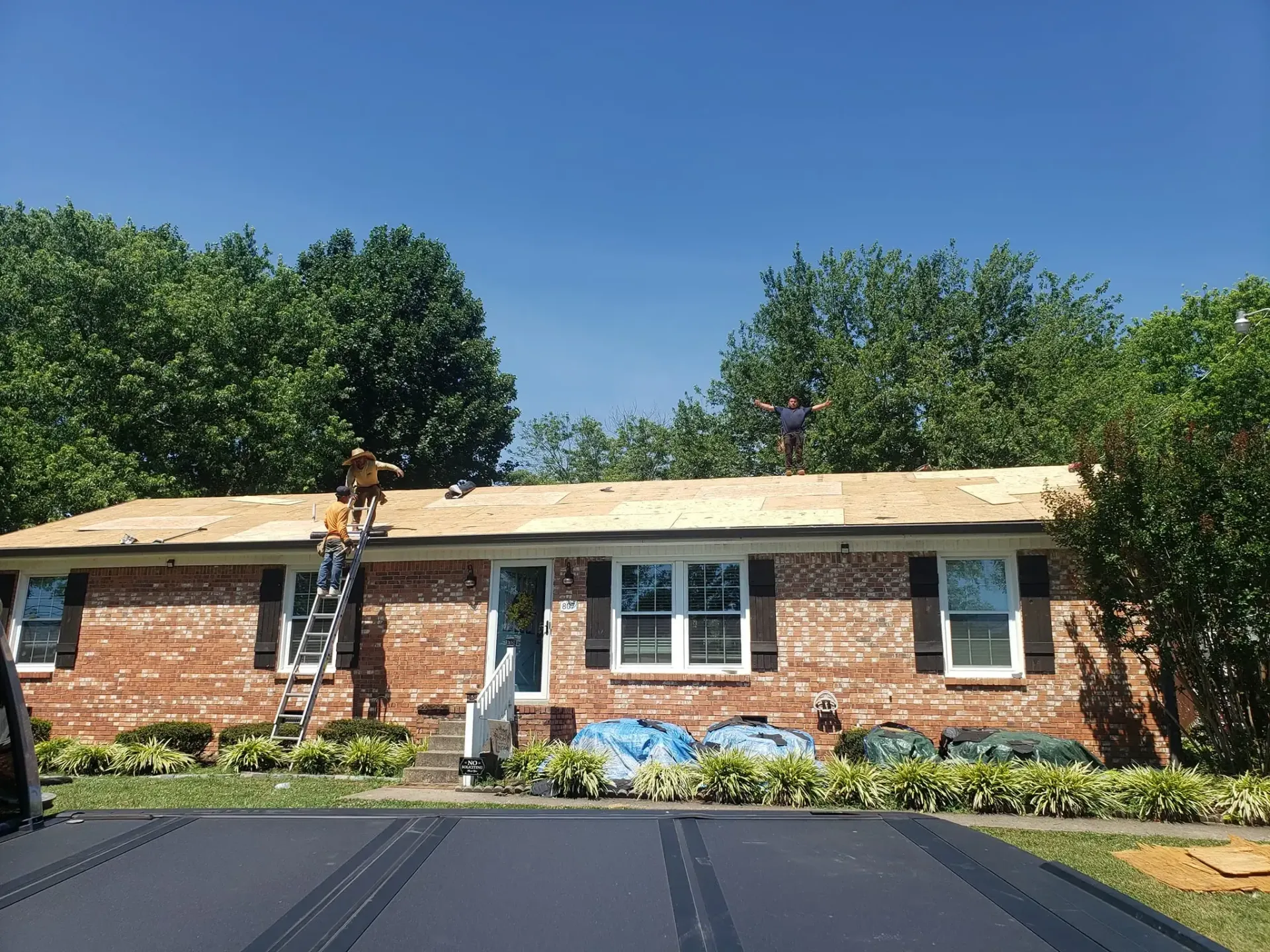 Roofers working on a brick house roof under a clear blue sky, surrounded by green trees.