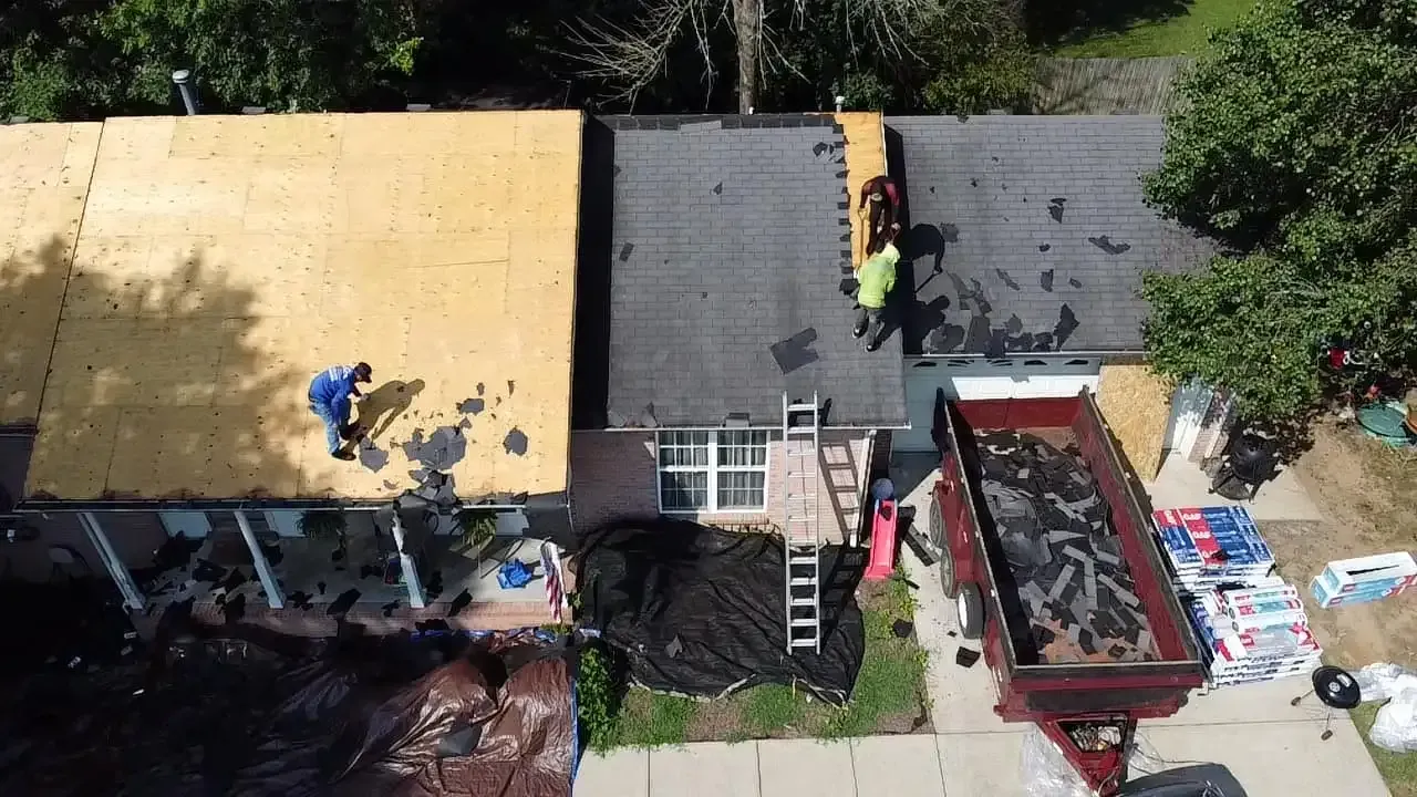 Roofers working on a house roof with exposed wood on one side, a dumpster, and debris below.