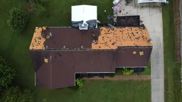 Aerial view of a house with workers removing shingles from the roof, showing exposed underlayment and debris.