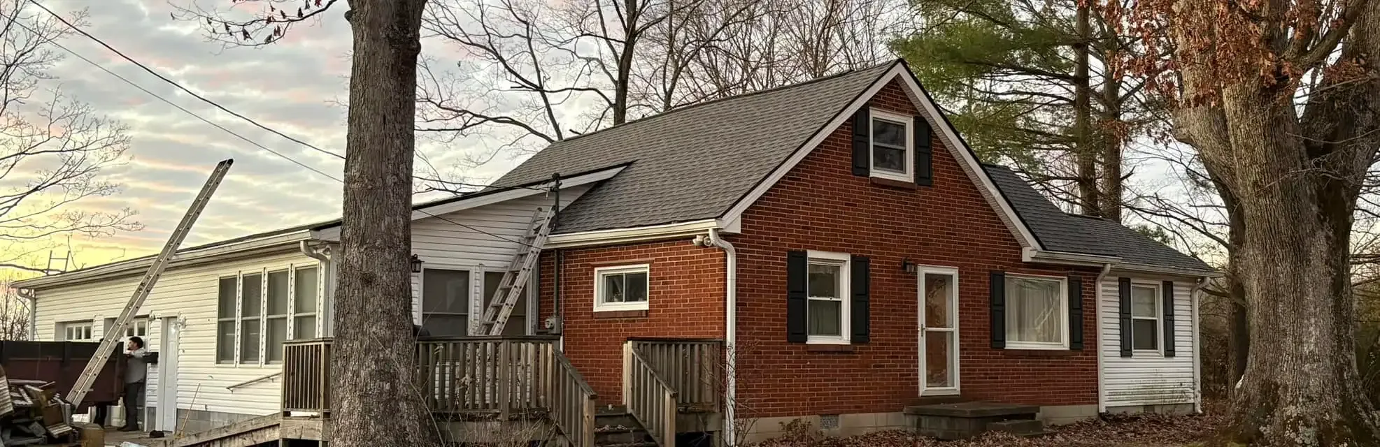 A two-story house with red brick facade, gray roof, and white siding. Trees surround the house.