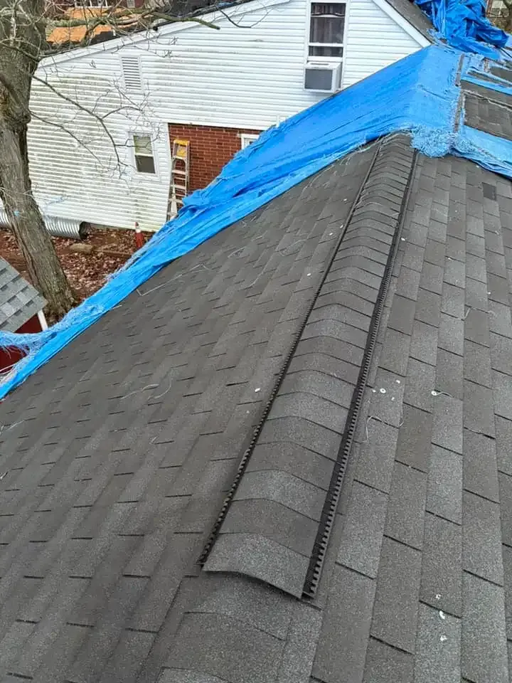 A shingled roof, partially covered with blue tarp, damaged. Building in the background.