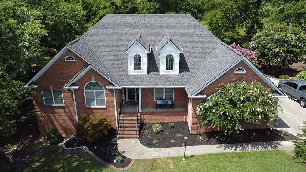 Red brick house with gray roof, dormers, and landscaping on a sunny day.