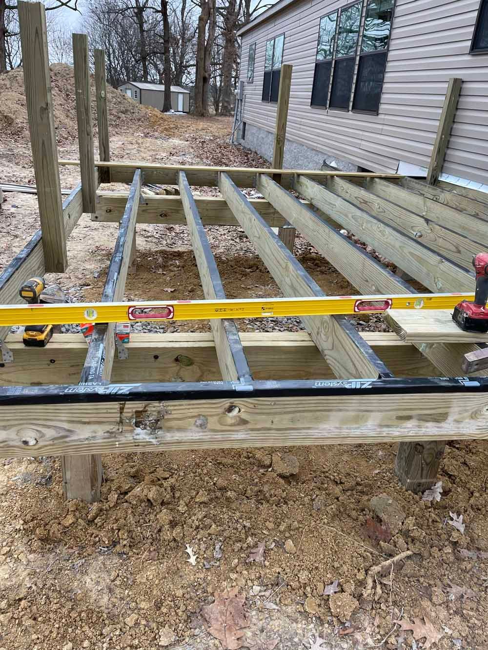 A wooden deck under construction with a level in use. Brown dirt surrounds.