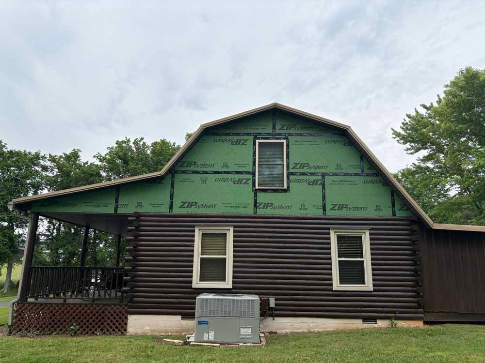 Cabin exterior under construction, green wrap on upper half, brown logs below, cloudy sky.