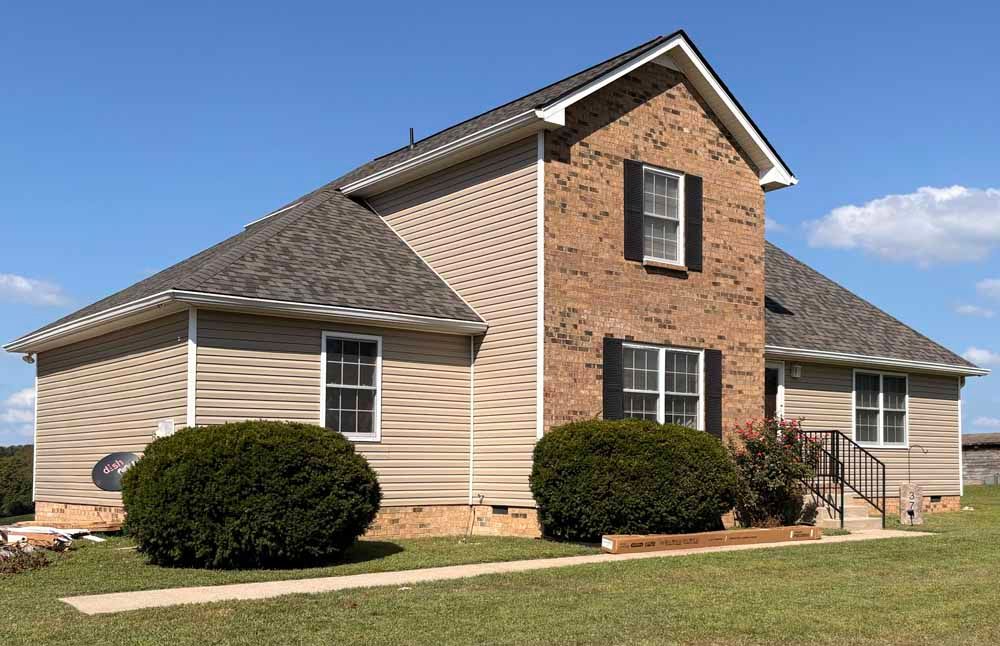 House with brick and beige siding, dark roof, green bushes, and blue sky.