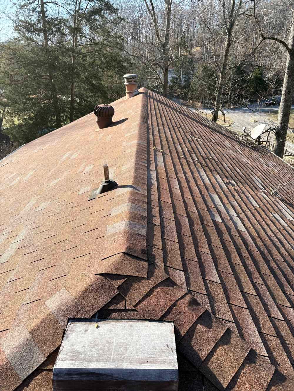 Brown asphalt shingle roof with chimney, vent, and deteriorating shingles. Sunlight illuminates the scene.
