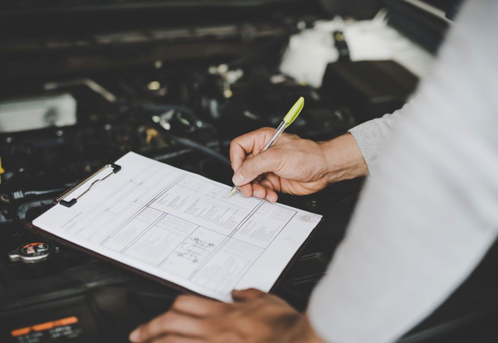 A Man Is Writing On A Clipboard In Front Of A Car Engine — Coffs Harbour Automotive In Coffs Harbour, NSW