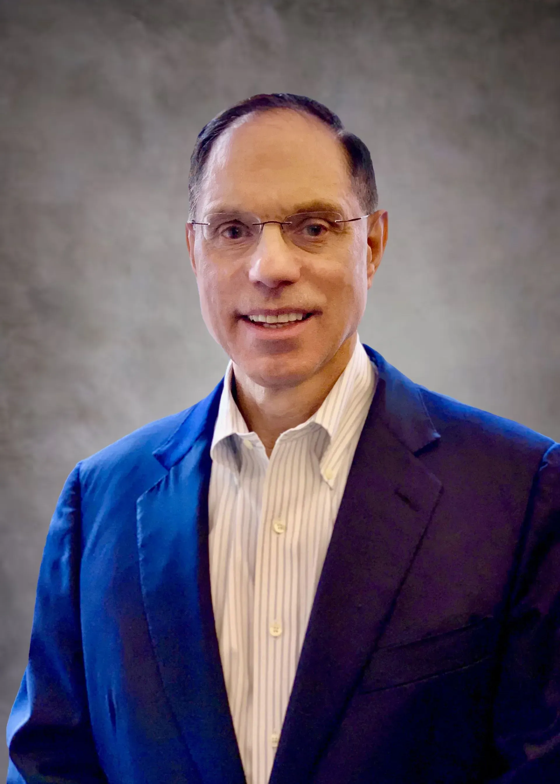 Man wearing glasses, blue blazer over a light pinstriped shirt, smiling, neutral background.