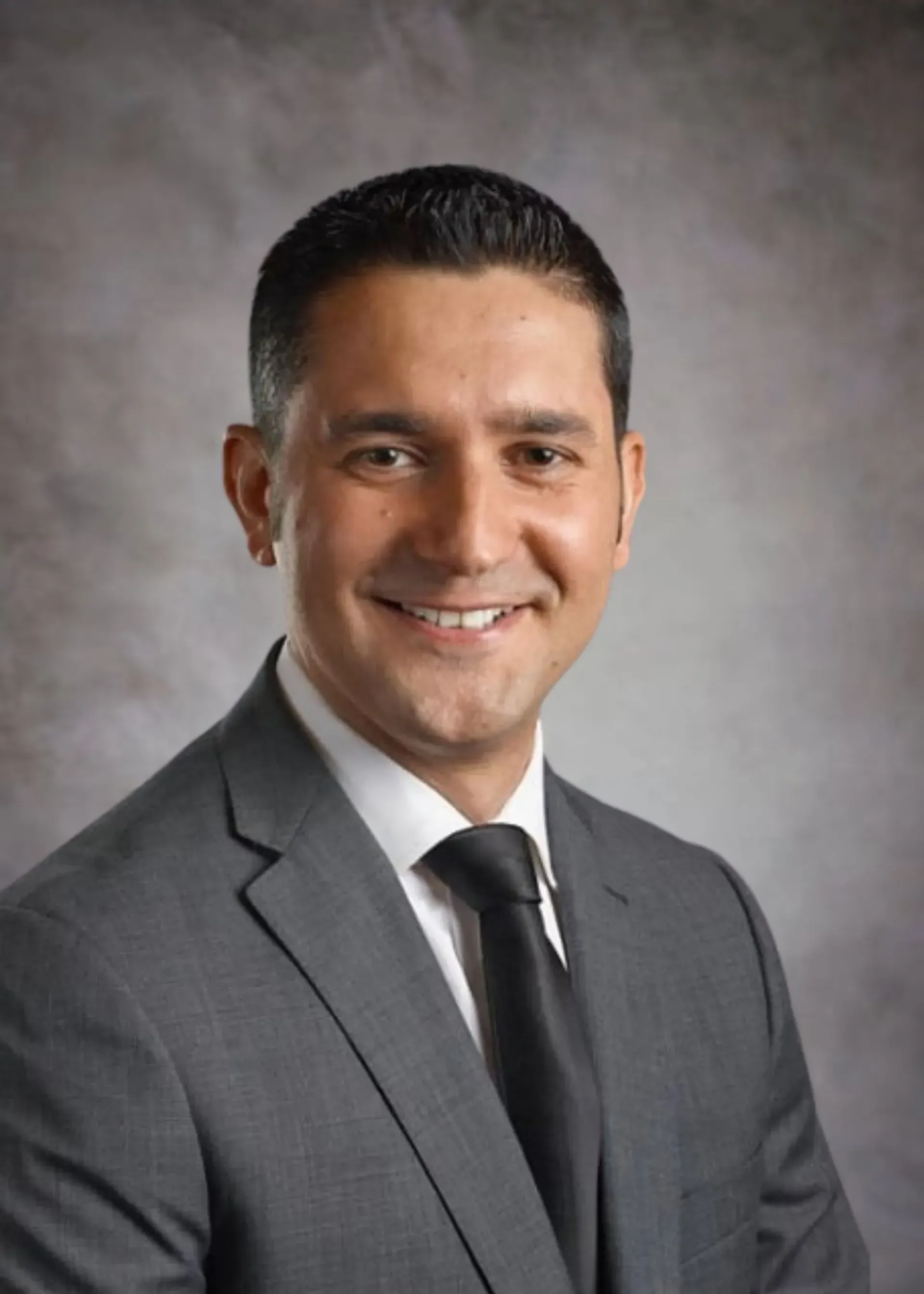 Man in gray suit and black tie, smiling against a mottled gray background.