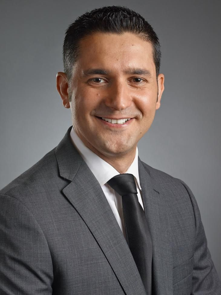 Man in gray suit and black tie, smiling against a mottled gray background.